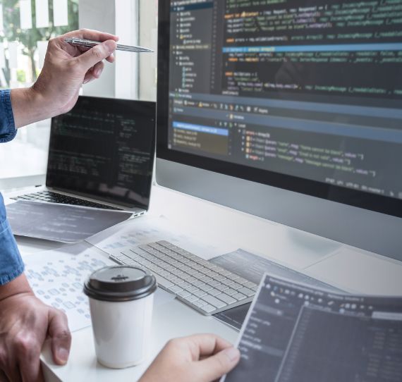 imagem de ecrãs de computador a correrem um script, mão com caneta a apontar para ecrã e mão de outra pessoa a segurar uma folha de papel com código
